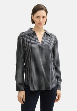 Vrouw in een losvallend, lang mouw, zwart-grijs geblokt shirt met een v-hals, gecombineerd met donkere broek.