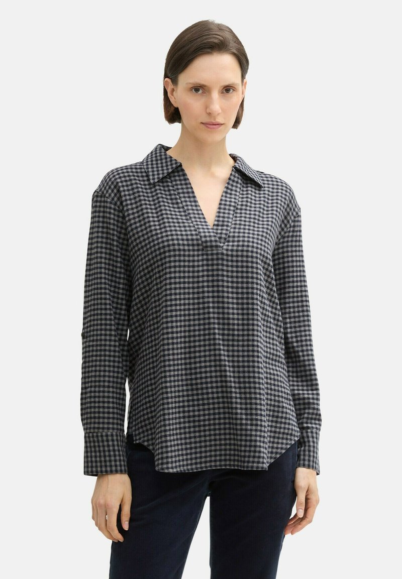 Femme portant une chemise à manches longues, ample, à carreaux noirs et gris avec un col en V, associée à un pantalon sombre.