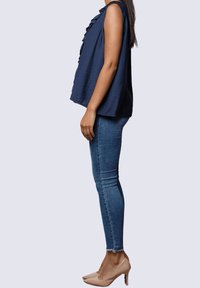 Top sin mangas color marino con volantes, combinado con jeans ajustados azules y zapatos de tacón nude, visto de lado. Textura de tela suave.