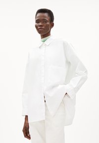 White oversized shirt made of cotton, featuring a button-down front, a single pocket, and a long hem with side slits.