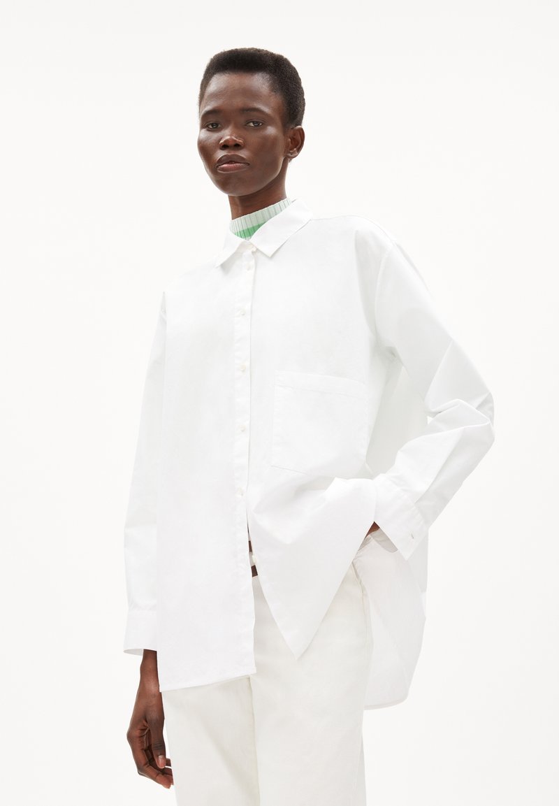 White oversized shirt made of cotton, featuring a button-down front, a single pocket, and a long hem with side slits.