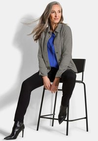 Grijze textuur blazer over een blauwe satijnen top, gecombineerd met zwarte slim-fit broek en zwarte puntige enklaarzen, zittend op een zwarte stoel.