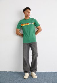 Grøn kortærmet t-shirt med en vandret rød, orange og gul stribe og "ellesse SPORT" logo. Båret med grå jeans og hvide sneakers.