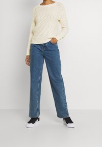 Gräddfärgad stickad tröja med ett strukturerat mönster, kombinerad med löst sittande blå denimjeans. Svarta sneakers med vita sulor och detaljer.