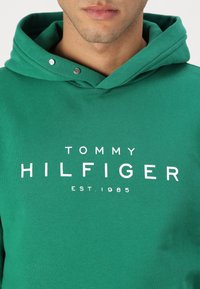 Zelená bavlněná mikina s kapsou klokankou, kapucí se šňůrkou a bílým tištěným logem: "TOMMY HILFIGER EST. 1985."