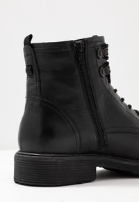 Svart läderankboots med en strukturerad yta, sidodragkedja och runda snörhål. Robust gummisula med subtila räfflor.