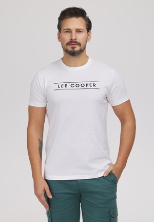 Mężczyzna z krótkimi ciemnymi włosami i brodą nosi białą koszulkę Lee Cooper oraz turkusowe spodnie typu cargo, stoi z jedną ręką w kieszeni na jednolitym tle.