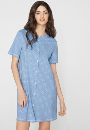 Joven mujer con cabello largo y oscuro que lleva un vestido azul claro de manga corta con botones y pequeños lunares blancos, de pie frente a un fondo liso.