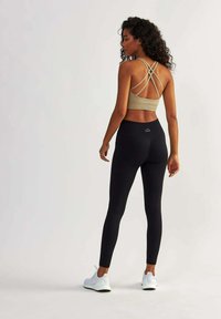 Soutien-gorge de sport tan avec des bretelles croisées, associé à des leggings noirs taille haute. Le modèle porte des chaussures de sport blanches. Texture de tissu lisse.