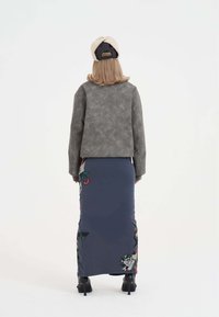 Giacca grigia con texture e schiena più corta, gonna a tubino blu con motivi floreali colorati e stivaletti neri con tacco.