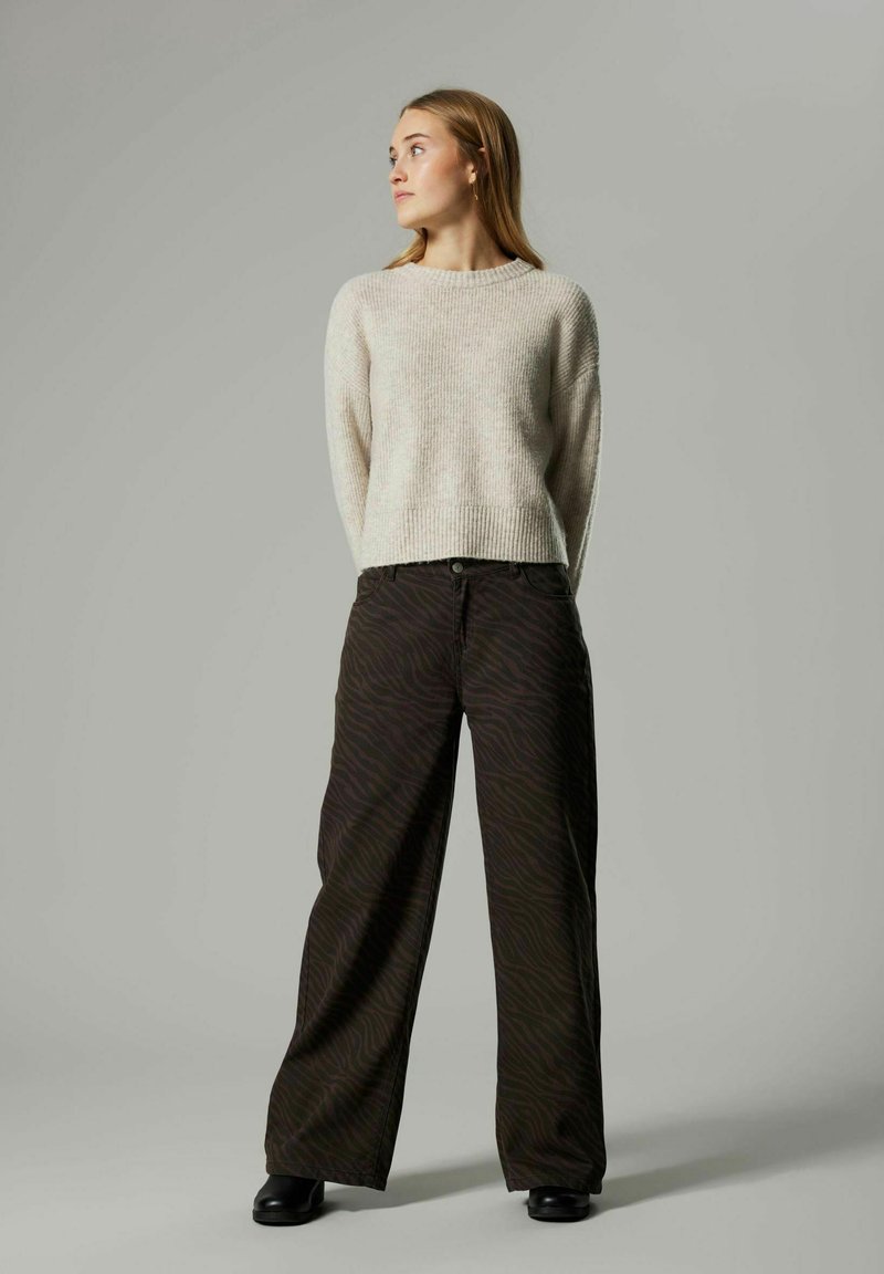 Femme se tenant avec les mains derrière le dos, portant un pull beige, un pantalon à larges pattes à motifs sombres et des chaussures noires, regardant sur le côté.