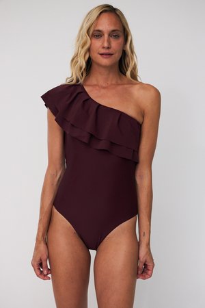Kobieta w ciemnofioletowym, asymetrycznym kostiumie kąpielowym z falbanką na ramieniu, stojąca na tle gładkiego jasnego tła.