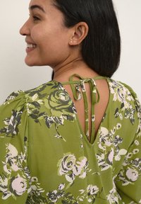 Grøn blomsterkjole med dyb V-ryg, der har pufærmer og en bindebåndslukning. Stoffet er letvægtigt og fremhæver et harmonisk blomsterprint.