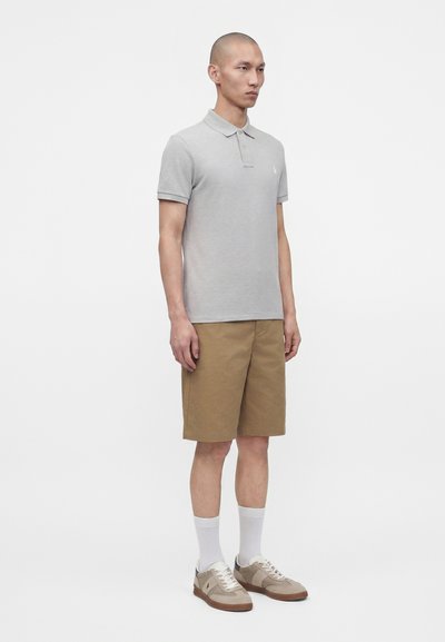 Homme debout face à l'avant portant un polo gris clair, un short marron jusqu'aux genoux, des chaussettes blanches et des baskets beiges sur fond blanc.