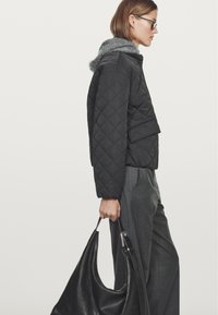 Black quilted jacket with a grey fur-lined hood, paired with grey trousers. The model holds a large black leather tote bag.