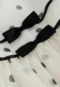 Tela blanca con lunares negros y grises, con dos lazos de satén negro y detalles fruncidos a lo largo del borde inferior.