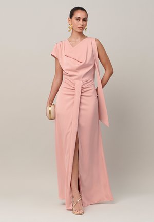 Vrouw in een roze jurk tot op de grond met een split, houdt een kleine beige clutch vast, draagt gouden oorbellen en gouden sandalen met bandjes.