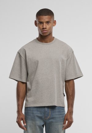 Rövid hajú férfi, laza szürke rövid ujjú ingben és kék farmernadrágban, egy sima világosszürke háttér előtt áll.