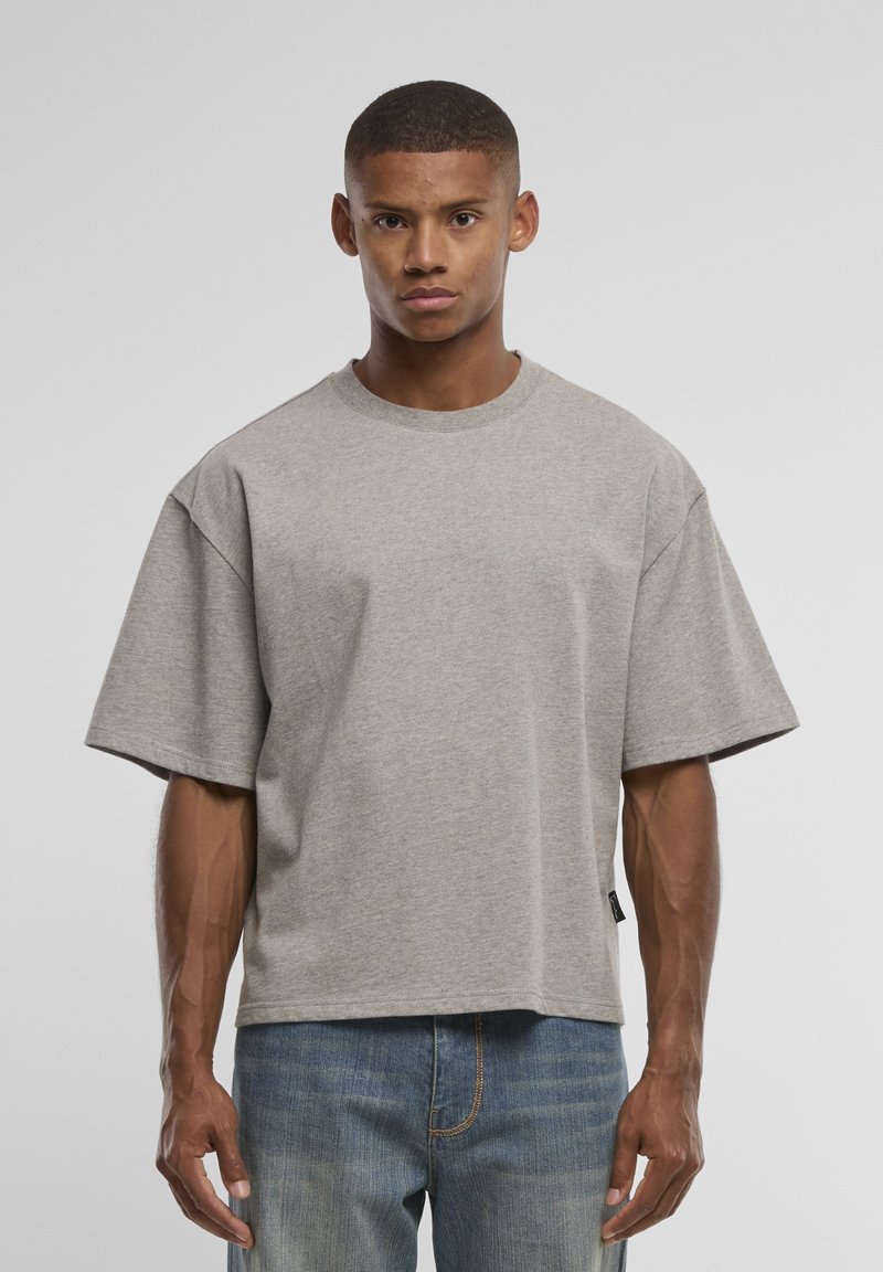 Homme aux cheveux courts portant une chemise ample grise à manches courtes et un jean bleu, debout devant un fond uni gris clair.