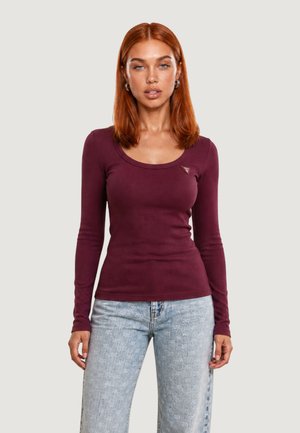 Mujer con cabello rojo liso lleva una camiseta morada ajustada de manga larga y vaqueros lavados azul claro sobre un fondo liso.
