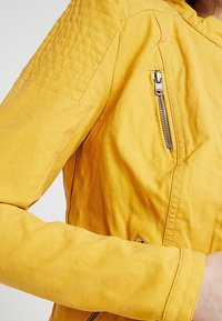 Giacca in pelle gialla con spalle testurizzate, tasche diagonali con zip e un design aderente, che mette in evidenza un materiale liscio e morbido.