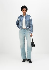 Jachetă bomber din satin bleu deschis cu accente albe, top alb din tricot, blugi bleu deschis cu talie înaltă și cizme cu toc negre, ținând o geantă mică neagră.