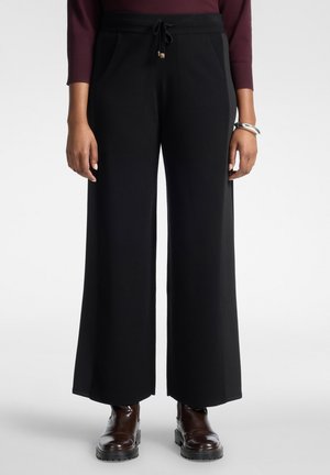 Pantalon noir à jambes larges avec taille à cordon, porté avec des bottines marron foncé et un haut bordeaux à manches longues sur une personne debout.
