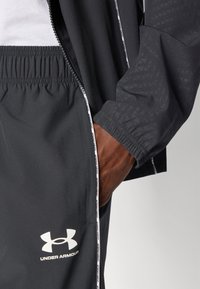 Jachetă athletică neagră cu un model geometric, fermoar în față, talie elastică și logo alb. Croi slim, țesătură ușoară.