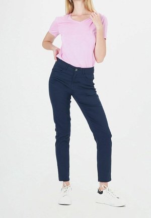 Femme portant un t-shirt rose clair à manches courtes, un pantalon bleu foncé ajusté et des baskets blanches, debout avec une main sur la hanche.