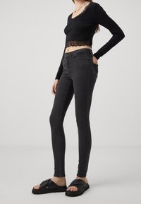 Mujer vestida con un top corto de encaje negro, jeans ajustados oscuros, sandalias negras de tiras cruzadas y un collar de perlas, de pie con una mano en el bolsillo.