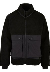 Giacca in pile nera con parte superiore a trama e parte inferiore in tessuto liscio. Presenta un colletto alto, chiusura frontale con zip e due tasche sul petto con bottone a pressione.