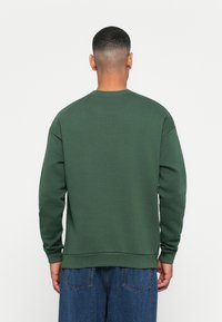 Felpa verde con scollo a girocollo, polsini e orlo a coste. Il tessuto ha una texture liscia, mostrata da dietro, abbinata a jeans di denim.