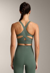 Zielony stanik sportowy z krzyżującym się tyłem, z wycięciami, wykonany z gładkiego, elastycznego materiału. Zawiera detal z logo na talii.
