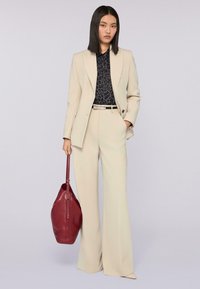Beige op maat gemaakt kostuum met een gestructureerde blazer en wijde broek, gecombineerd met een zwart met patroon overhemd en een dieprode leren handtas.