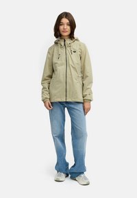 Lys beige lett jakke med hette, frontfyse, og snøring; paret med lyseblå rette jeans og grå joggesko.