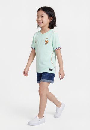 Νεαρό παιδί που περπατά, φορώντας ανοιχτό πράσινο αθλητικό μπλουζάκι με λογότυπο κόκορα, τζιν σορτς και λευκά παπούτσια χωρίς κορδόνια με φόντο απλό λευκό.