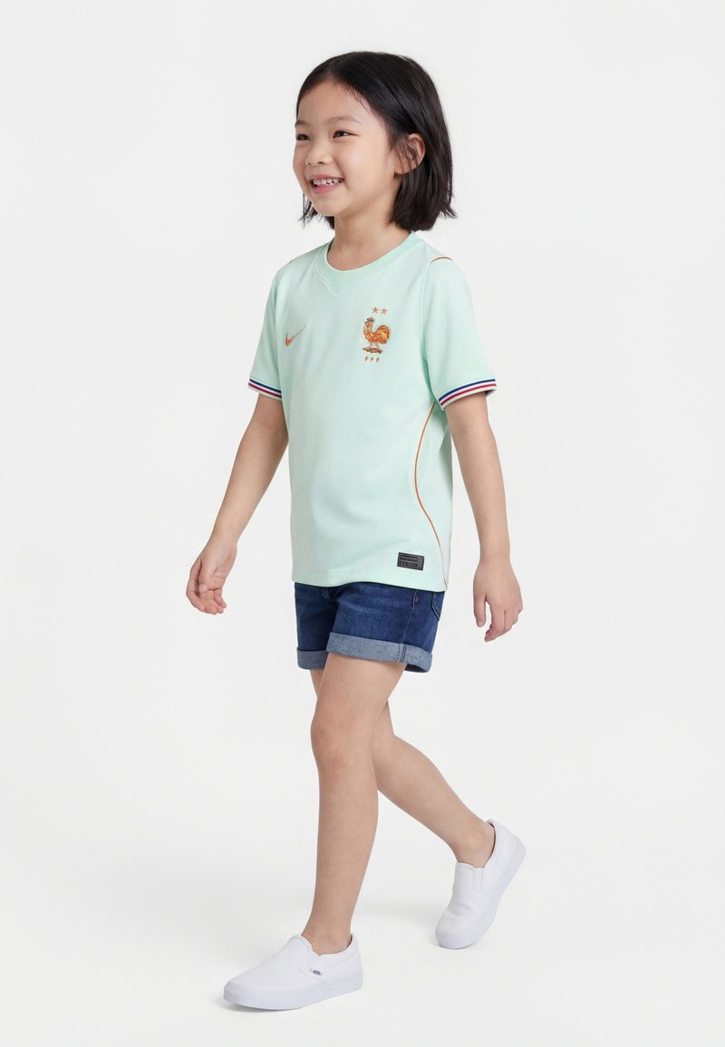 Νεαρό παιδί που περπατά, φορώντας ανοιχτό πράσινο αθλητικό μπλουζάκι με λογότυπο κόκορα, τζιν σορτς και λευκά παπούτσια χωρίς κορδόνια με φόντο απλό λευκό.