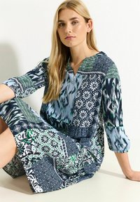 Vestido de manga larga con una mezcla de patrones en azul, verde y negro, con escote en V y tejido texturizado. Diseño de múltiples patrones en toda la prenda.