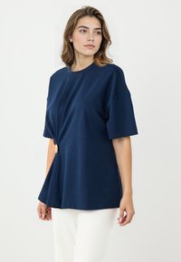 Marineblaue T-Shirt mit kurzen Ärmeln, rundem Halsausschnitt und einem Twist-Detail an der Vorderseite, das einen goldenen Knopfakzent an der Seite bietet.