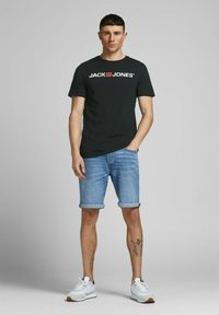 Schwarzes T-Shirt mit kurzen Ärmeln und "JACK & JONES"-Logo, kombiniert mit umgeschlagenen Denim-Shorts und weißen Sportschuhen. Sauberer, lässiger Look.