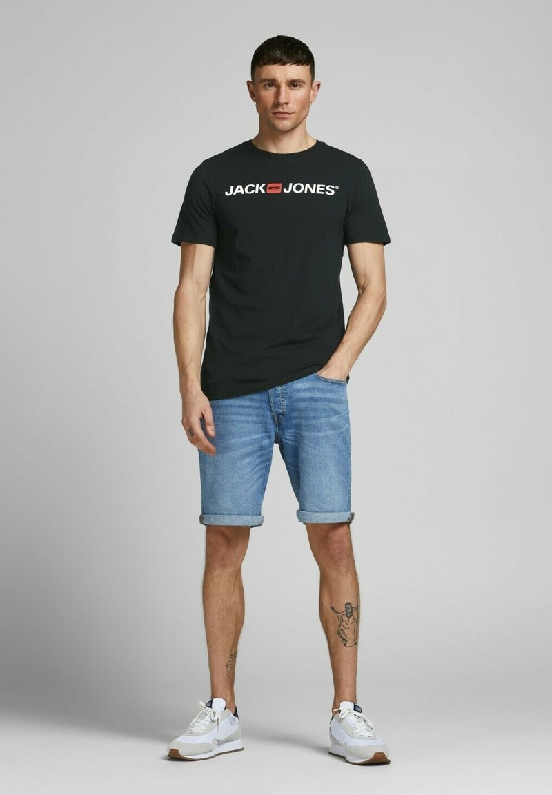 Schwarzes T-Shirt mit kurzen Ärmeln und "JACK & JONES"-Logo, kombiniert mit umgeschlagenen Denim-Shorts und weißen Sportschuhen. Sauberer, lässiger Look.