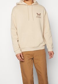 Sudadera con capucha beige con bolsillo canguro, capucha con cordón y gráfico de mariposa marrón con el texto "La abundancia es una mentalidad" en el pecho.