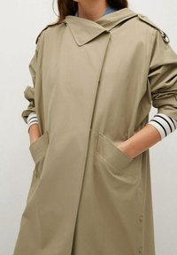 Imperméable beige avec grand col, fermeture à boutons et poches latérales. Texture douce et coupe décontractée avec manches rayées retroussées.