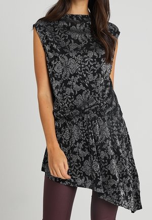 Kobieta nosząca czarną tunikę bez rękawów z srebrnym kwiatowym wzorem i asymetrycznym dołem na ciemnofioletowych legginsach.