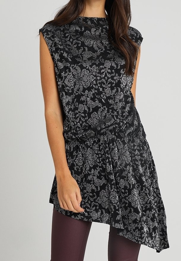 Femme portant une tunique noire sans manches avec un motif floral argenté et un ourlet asymétrique sur un legging violet foncé.