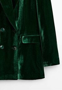 Grön sammetsblazer med dubbelknäppt design, belagd med mörka knappar, notch-krage och en enda bröstficka.