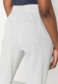 Zbliżenie osoby noszącej jasnoszare spodnie o fakturze, z elastycznym pasem i naszywanymi tylnymi kieszeniami, stojącej z prawą ręką luźno opuszczoną.