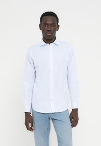 Άσπρο πουκάμισο με μακριά μανίκια και κουμπιά, με διακριτικό σχέδιο, που διαθέτει μυτερό γιακά και ίσια ραφή, φορεμένο με ανοιχτόχρωμο μπλε τζιν.