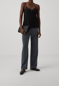 Vrouw in een zwart met kant afgewerkt hemd, grijze wijde broek, zwarte puntige flats en met een zwart geembosst clutch.