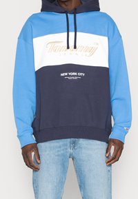 Hoodie color-blocking in blu, bianco e navy. Presenta il logo "Tommy Jeans" in scrittura e il testo "NEW YORK CITY". Tessuto morbido e texturizzato.